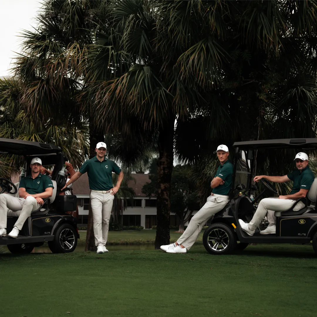 Four golfers in green shirts and white pants pose with golf carts on a grassy area with palm trees in the background.