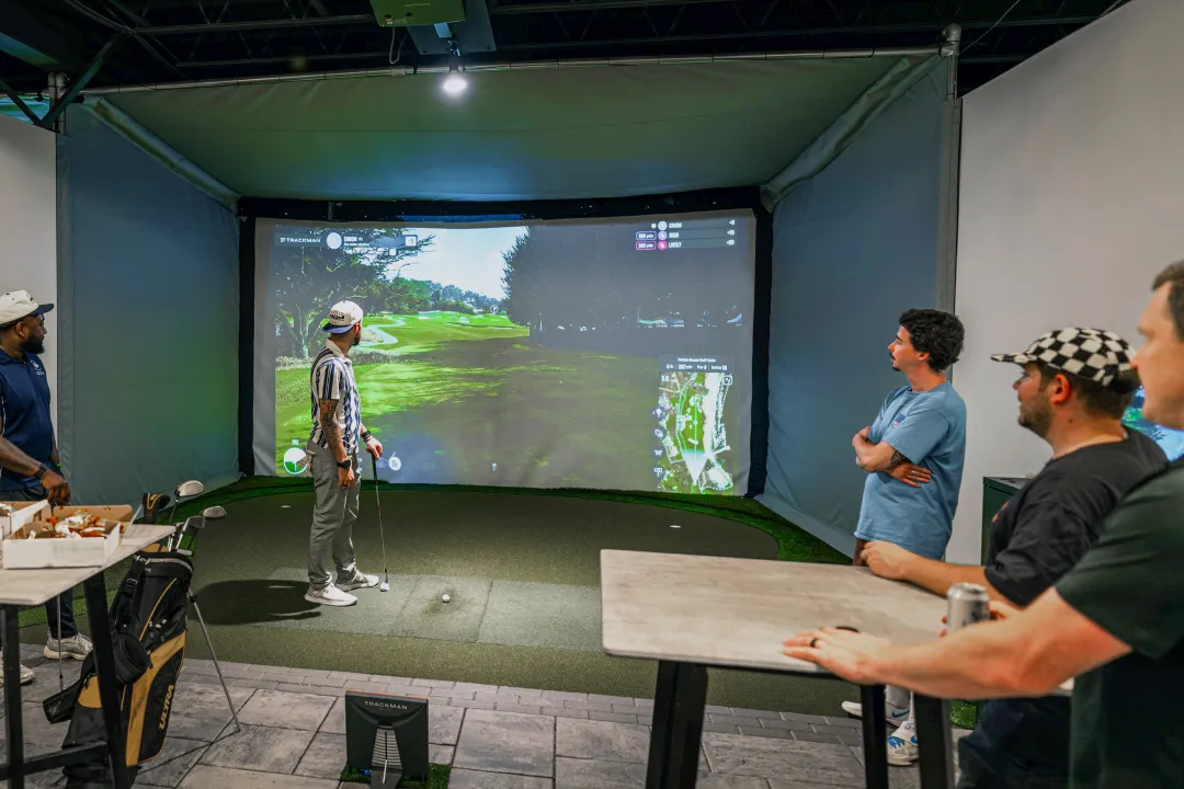 A group of people in an indoor golf simulator, watching as one prepares to hit a golf ball. A virtual golf course is projected on a large screen.