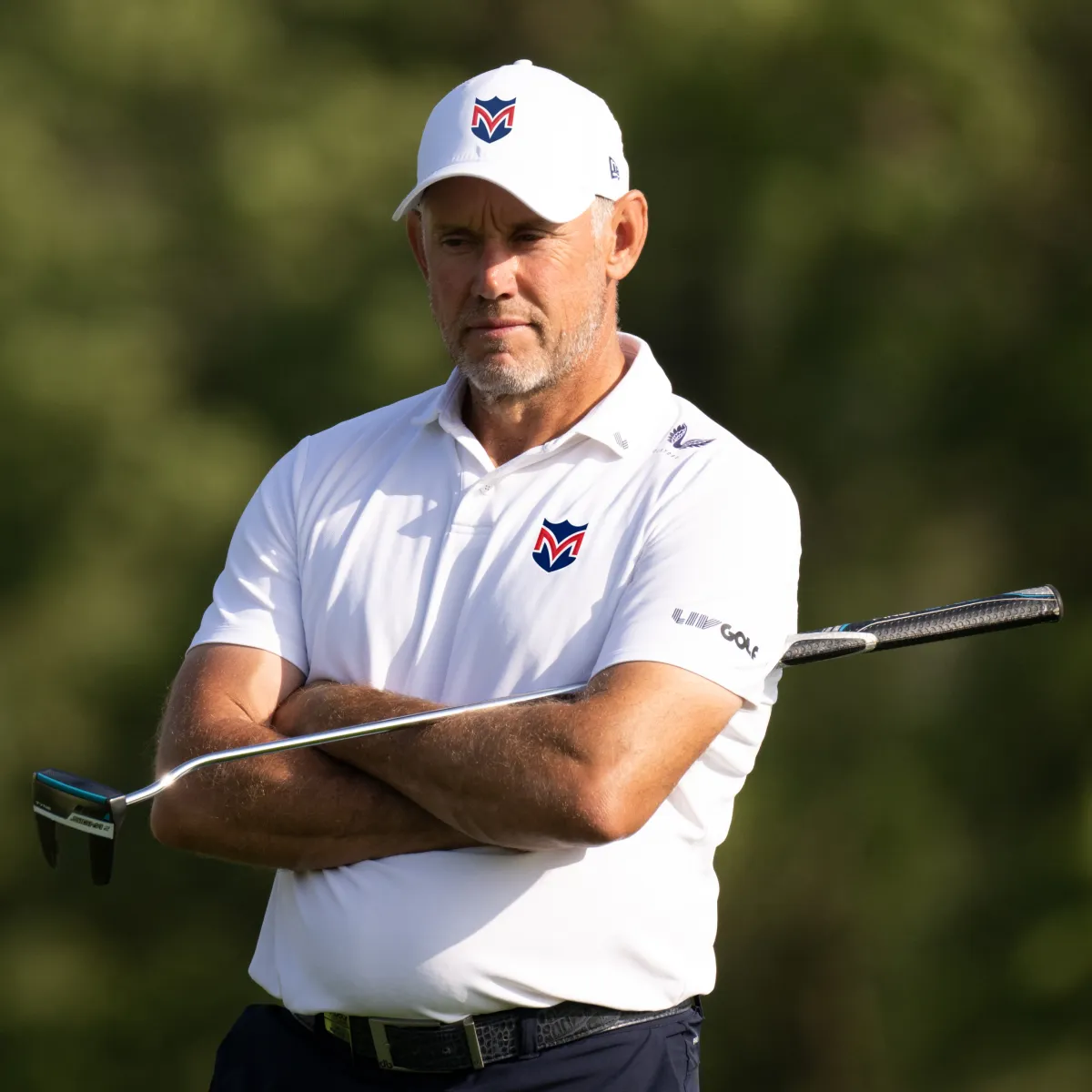 A man in a white hat and polo shirt stands with arms crossed, holding a golf club against his chest, outdoors on a golf course.