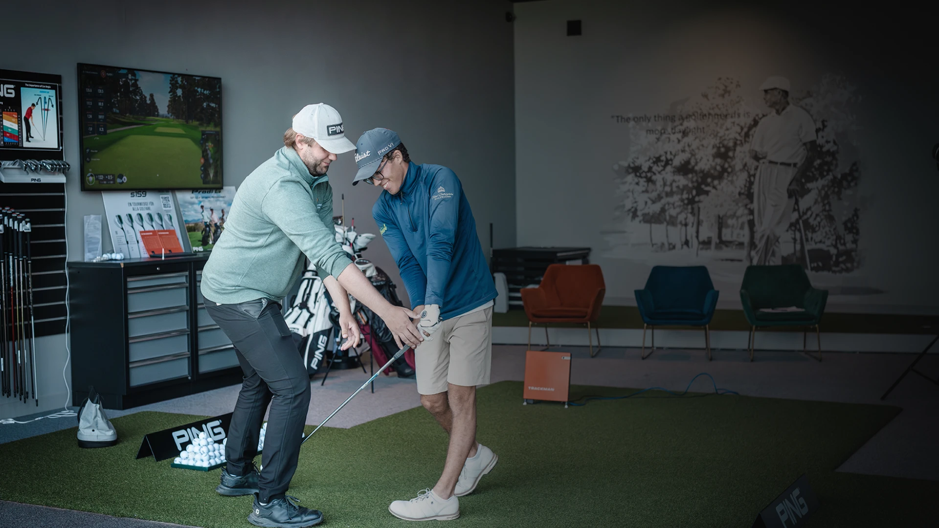 Two people in a golf store: one instructing the other on a swing. Golf equipment displayed in the background.