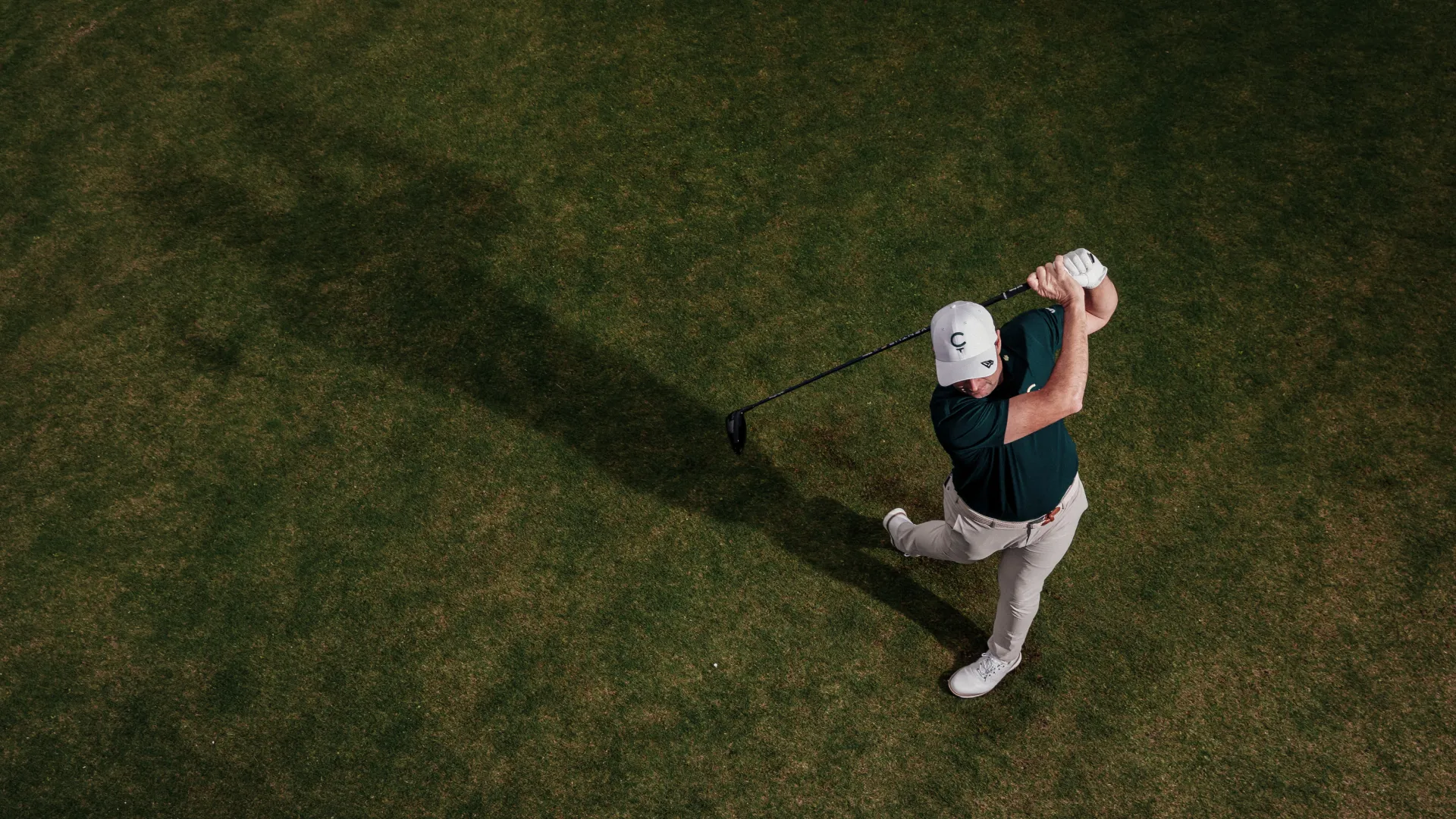 Golfer in a white cap swings a club on a green field, casting a long shadow. The image is taken from above, highlighting the dynamic motion.