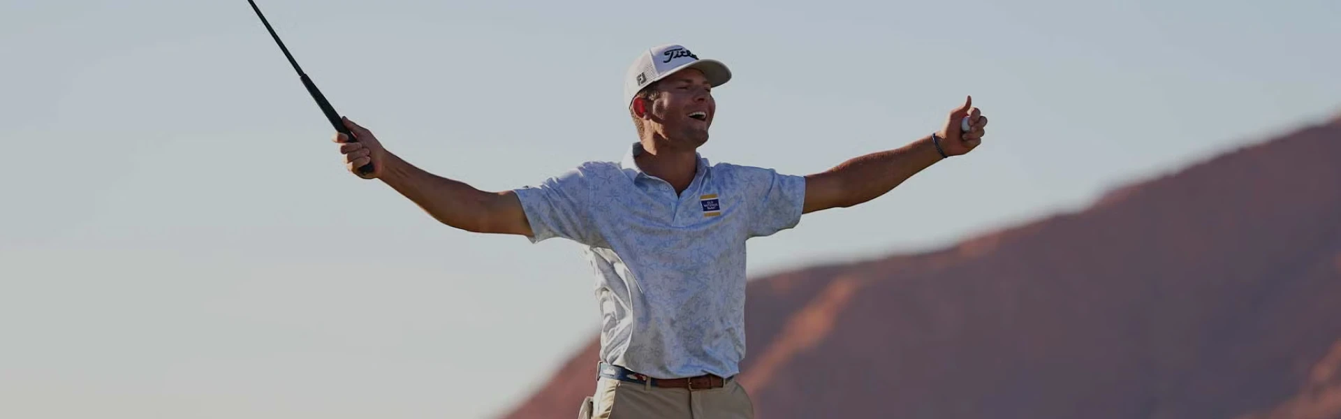 Golfer in a light blue shirt and white cap celebrates with arms outstretched, holding a golf club, against a backdrop of hills and clear sky.