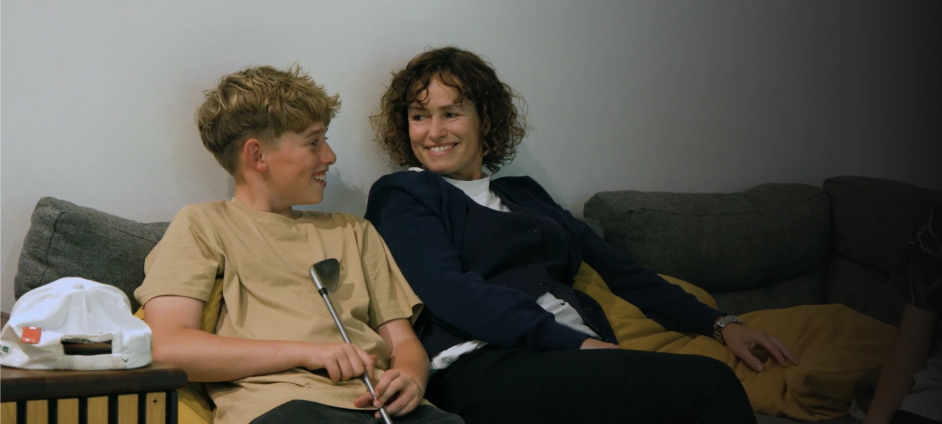 A woman and a young boy sit on a couch, smiling at each other. The boy holds a golf club, and a cap is beside him.