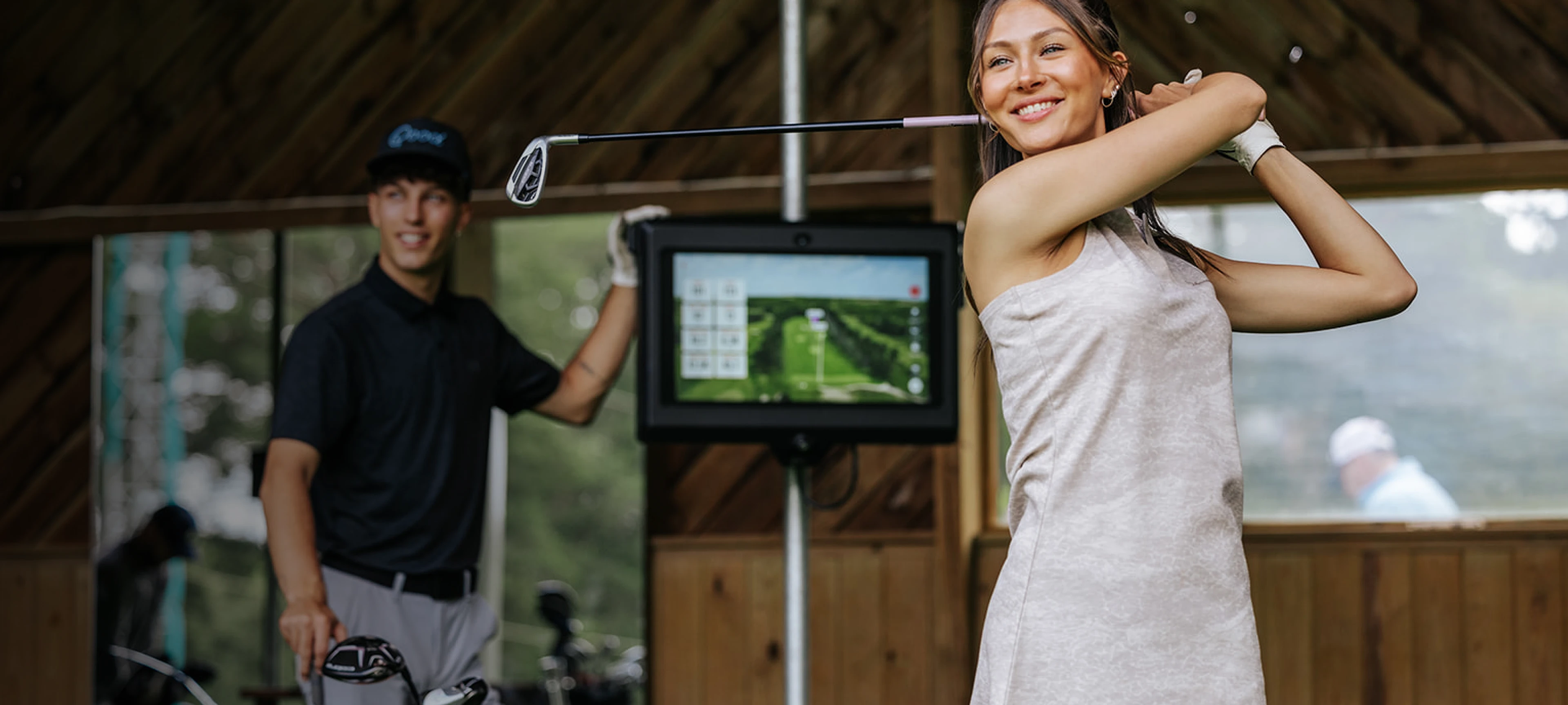 Woman swings a golf club inside a range, smiling, while a man watches in the background next to a screen displaying golf data.