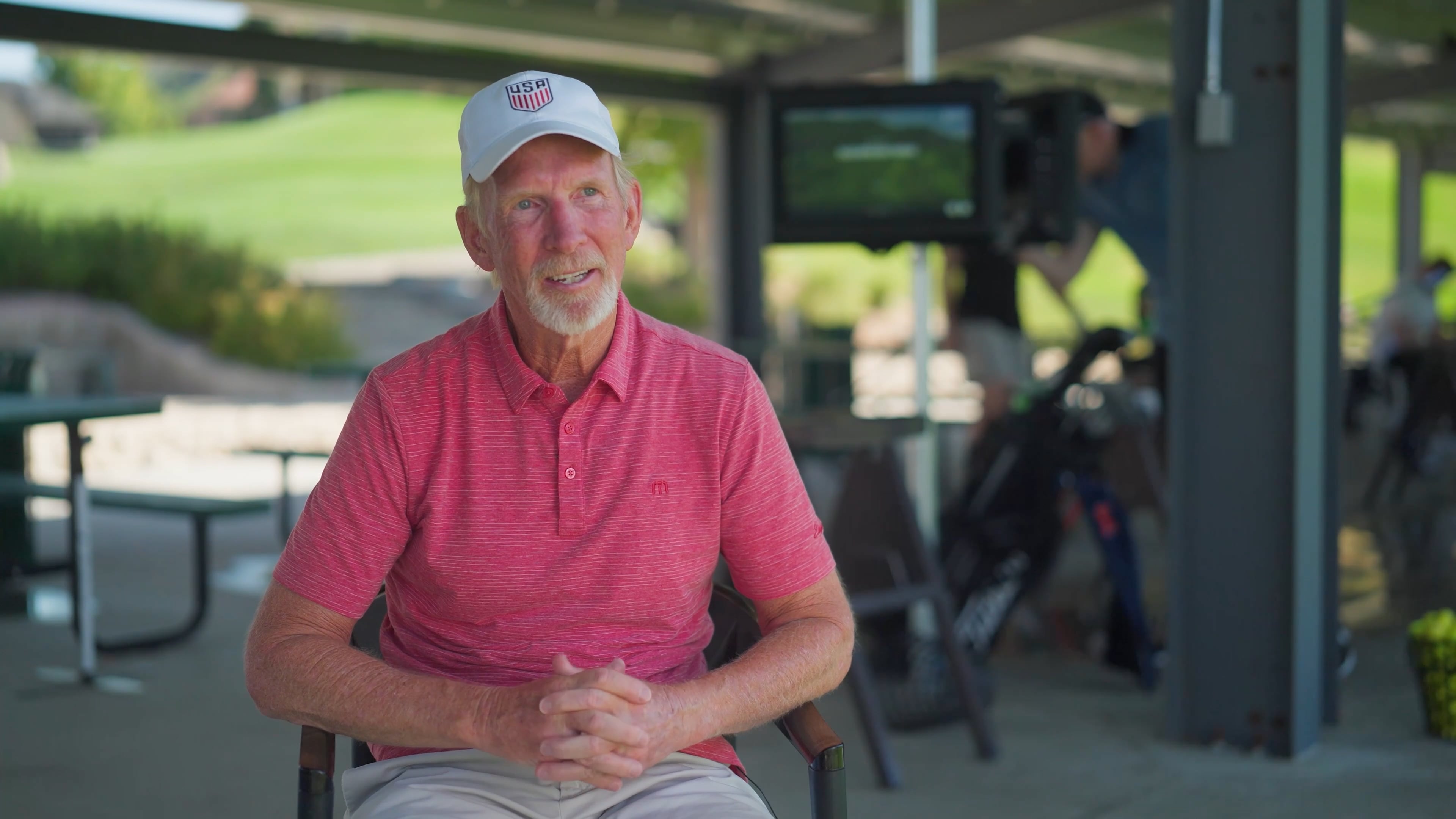 Elderly man in a red shirt and white cap sitting in a golf driving range, smiling, with golf bags and a TV screen in the background.