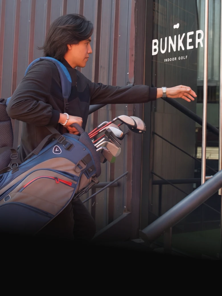 Person entering an indoor golf venue carrying a golf bag over their shoulder. The entrance features "BUNKER" signage.