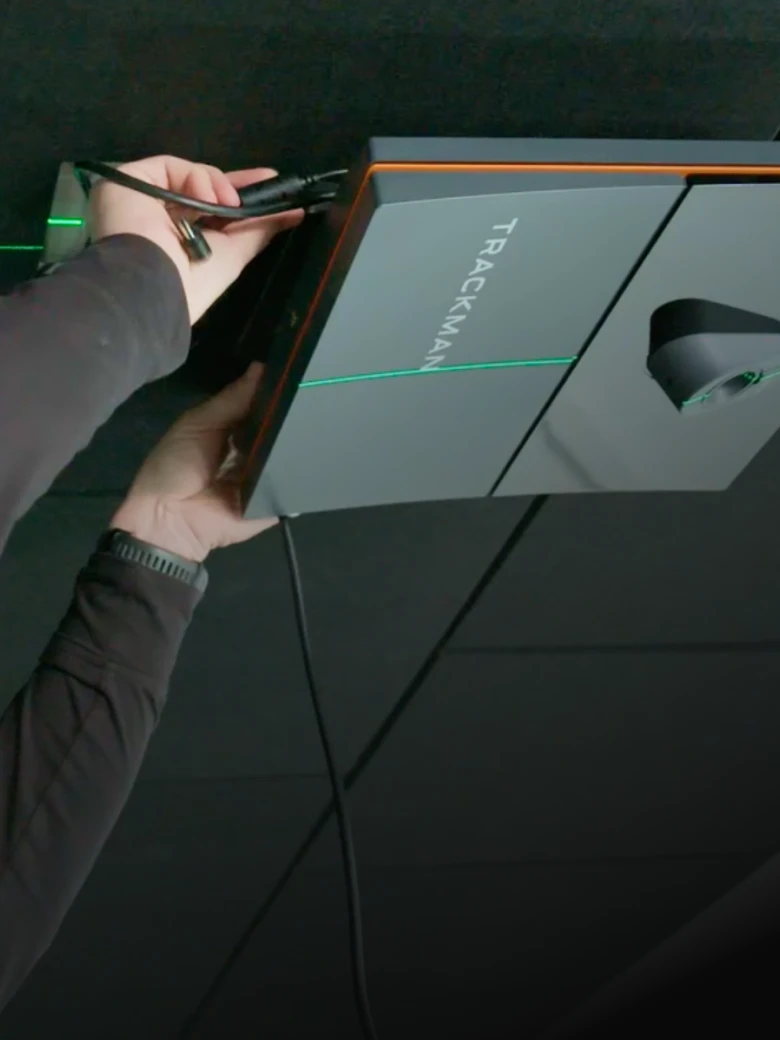 Hands adjusting a ceiling-mounted TrackMan device, with wires connected, against a dark background.