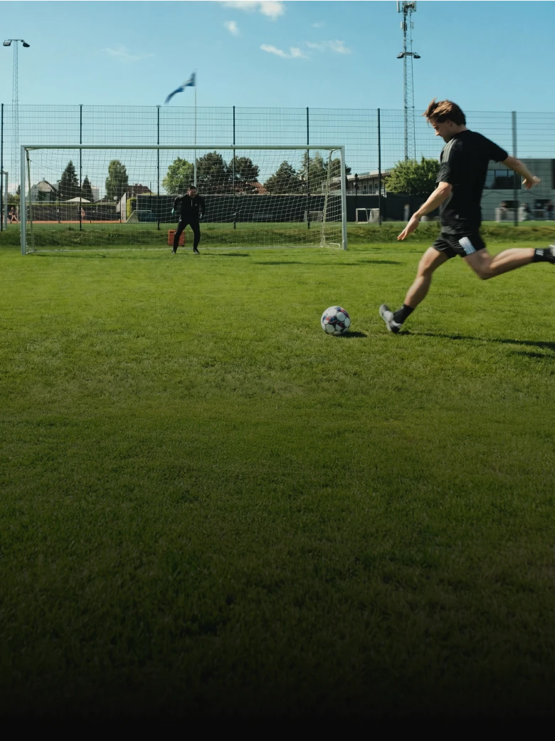 A soccer player in black attire prepares to kick a ball towards a goalkeeper standing in front of a goal on a grassy field.