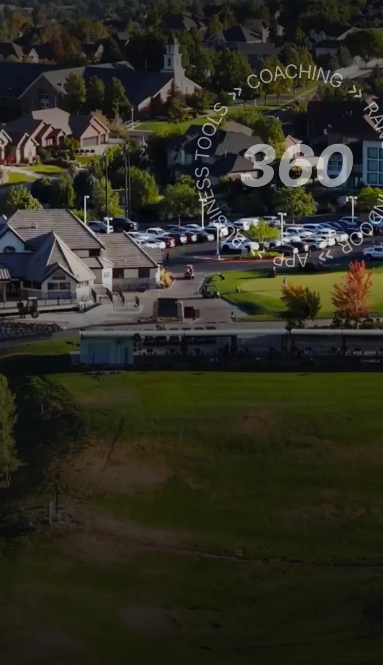 Aerial view of a suburban area with houses, a parking lot, and a grassy field. Trees with autumn foliage are visible.