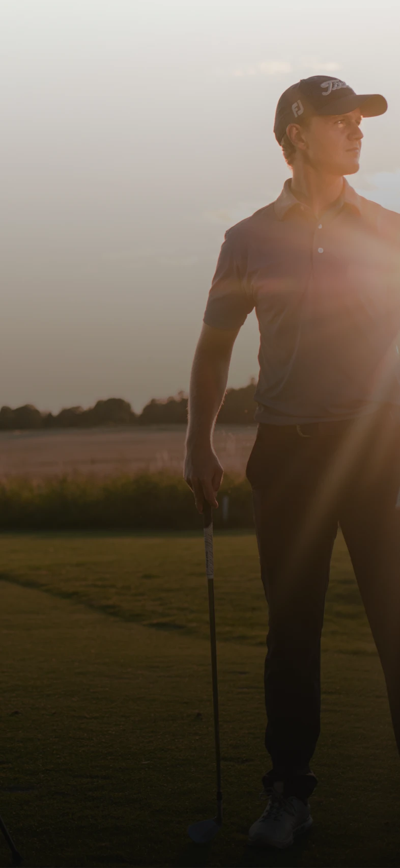 Golfer standing on a sunlit course holding a club, with a golf bag nearby. Sun is setting on the horizon, creating a warm glow.