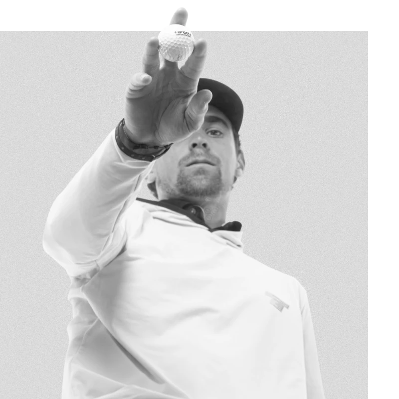 Person in a cap and jacket reaches forward to display a golf ball toward the camera, set against a plain background.