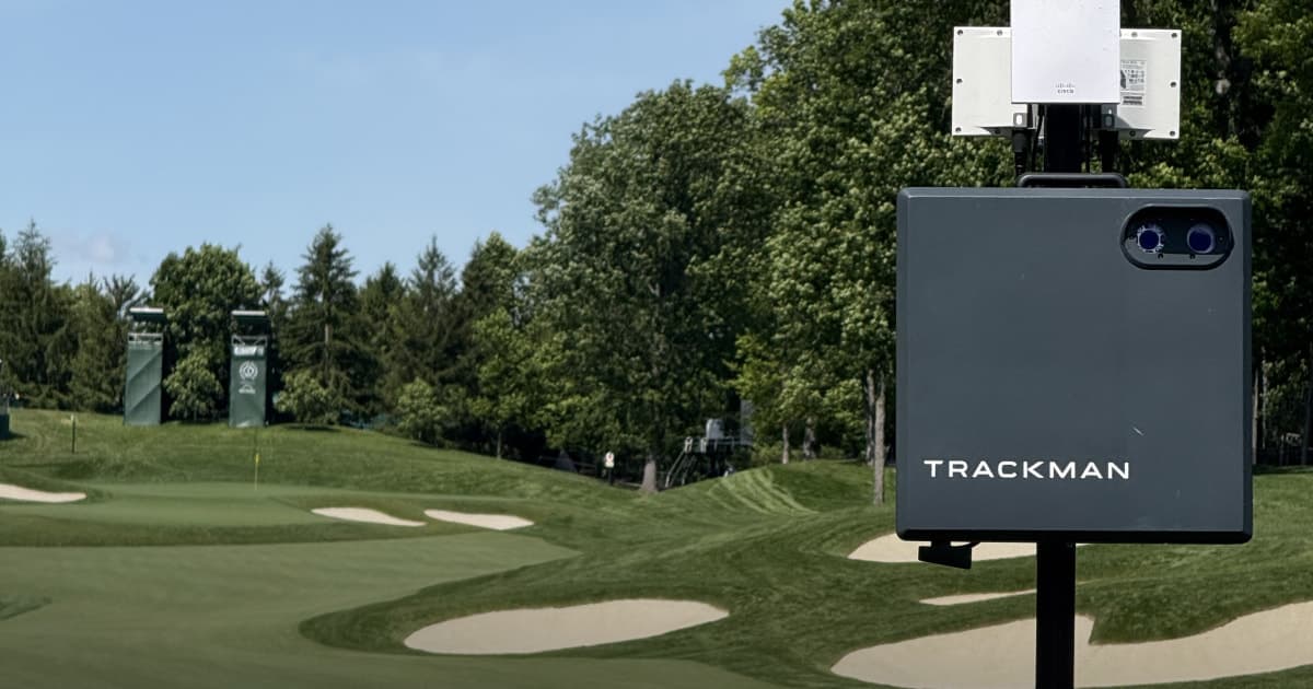 TrackMan golf radar device set up on a lush golf course with sand bunkers and trees in the background under a clear blue sky.