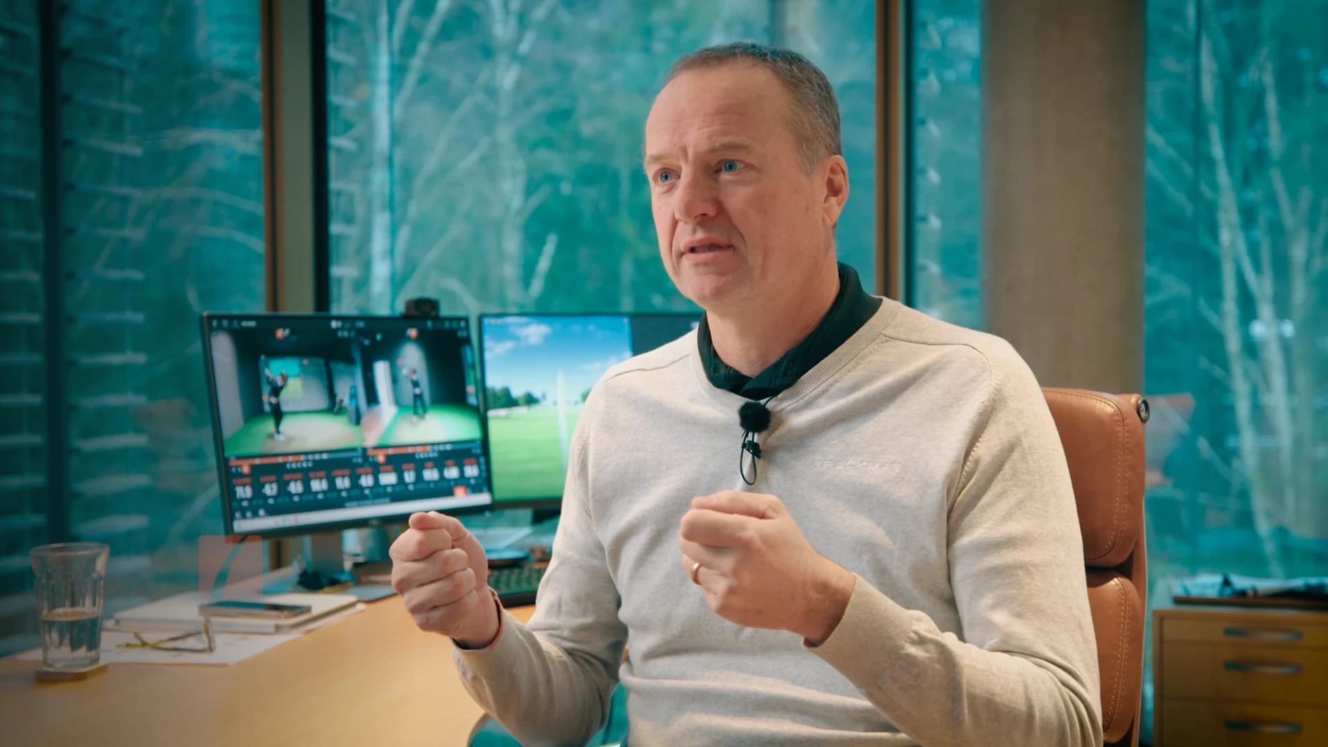 A man in a light sweater gestures while speaking in an office with large windows. Two computer monitors display golf-related content behind him.