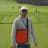 Brandon La Rosa, wearing a cap and sunglasses, stands on a golf course holding an orange case, with others in the background.
