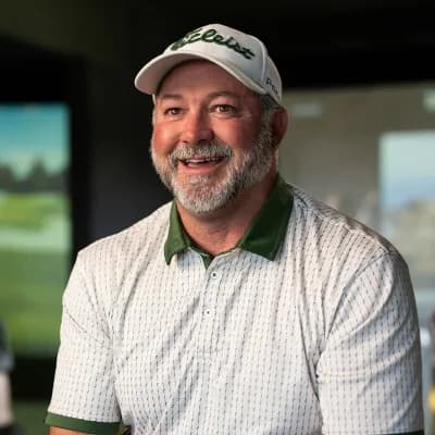 Man in a golf cap and polo shirt smiling indoors, with a blurred background displaying a golf course.