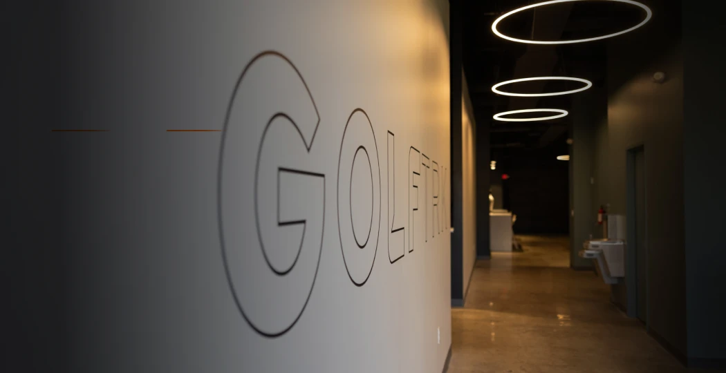 Dimly lit hallway with the word "GOLFTRIX" on the wall, featuring circular ceiling lights leading towards a reception area.