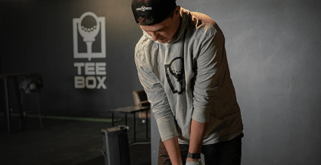A person in a gray sweatshirt and black cap is setting up to putt a golf ball indoors, with a "TEE BOX" logo on the wall behind.