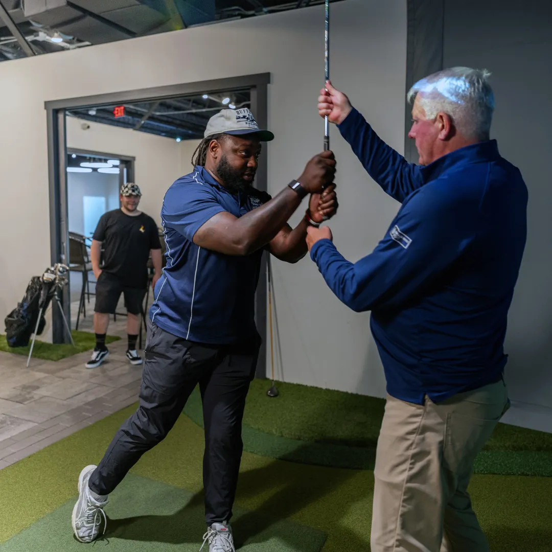 Two men practicing a golf swing indoors, with one man guiding the other's hands on a golf club. A third person watches in the background.