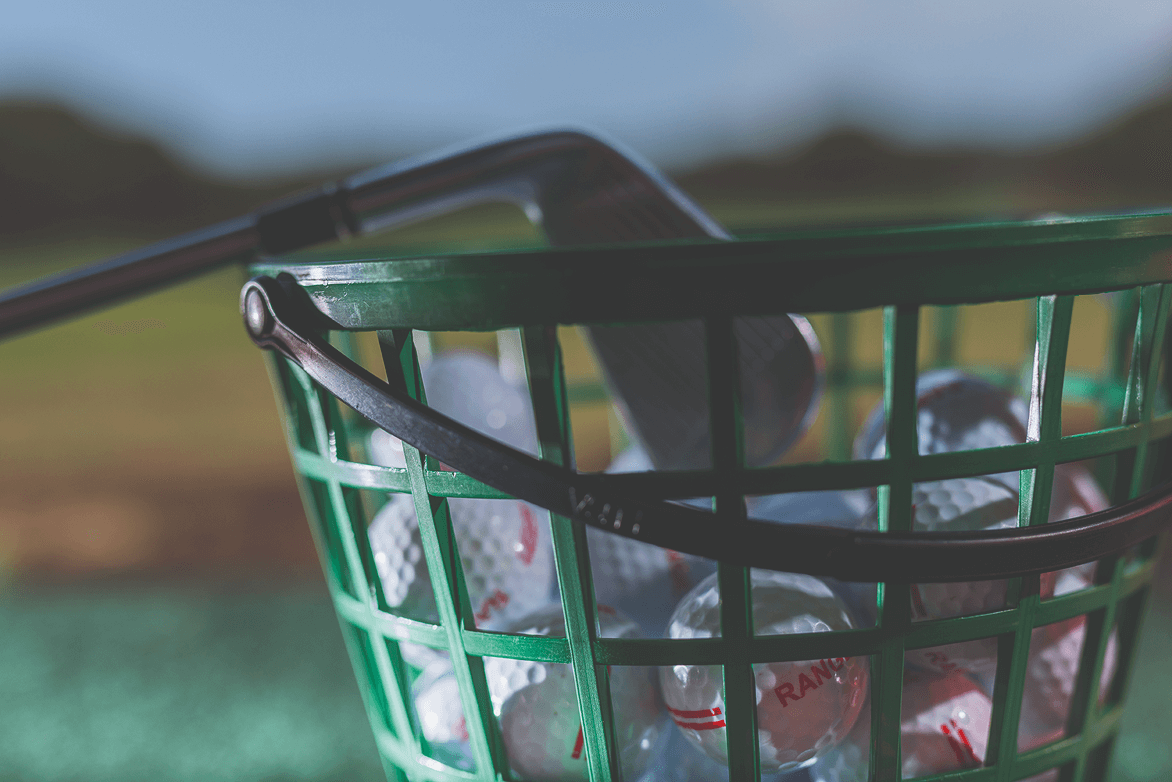 A green basket filled with golf balls and a golf club against a blurred outdoor background.