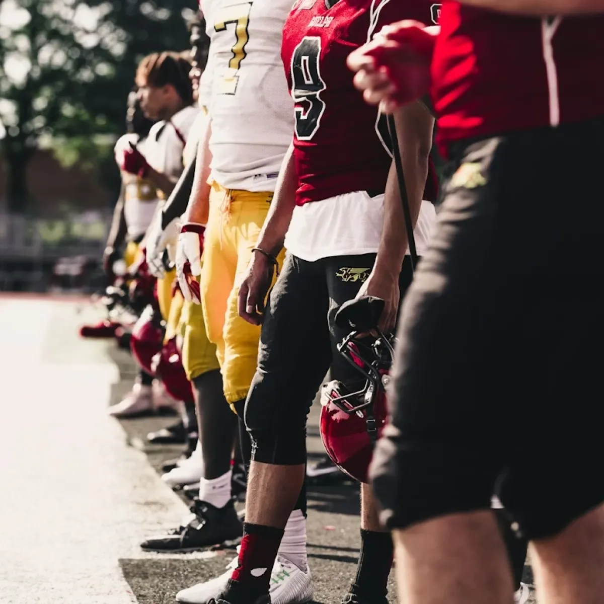 Football players lined up on the sideline, wearing red and yellow uniforms and holding helmets.