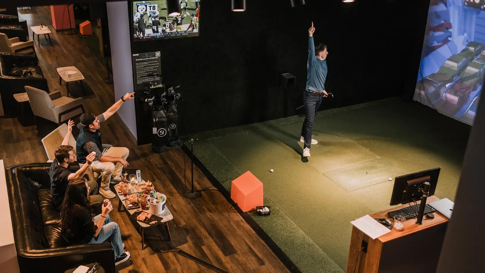 A person plays a golf simulation game indoors while friends watch from a couch, surrounded by snacks and drinks.