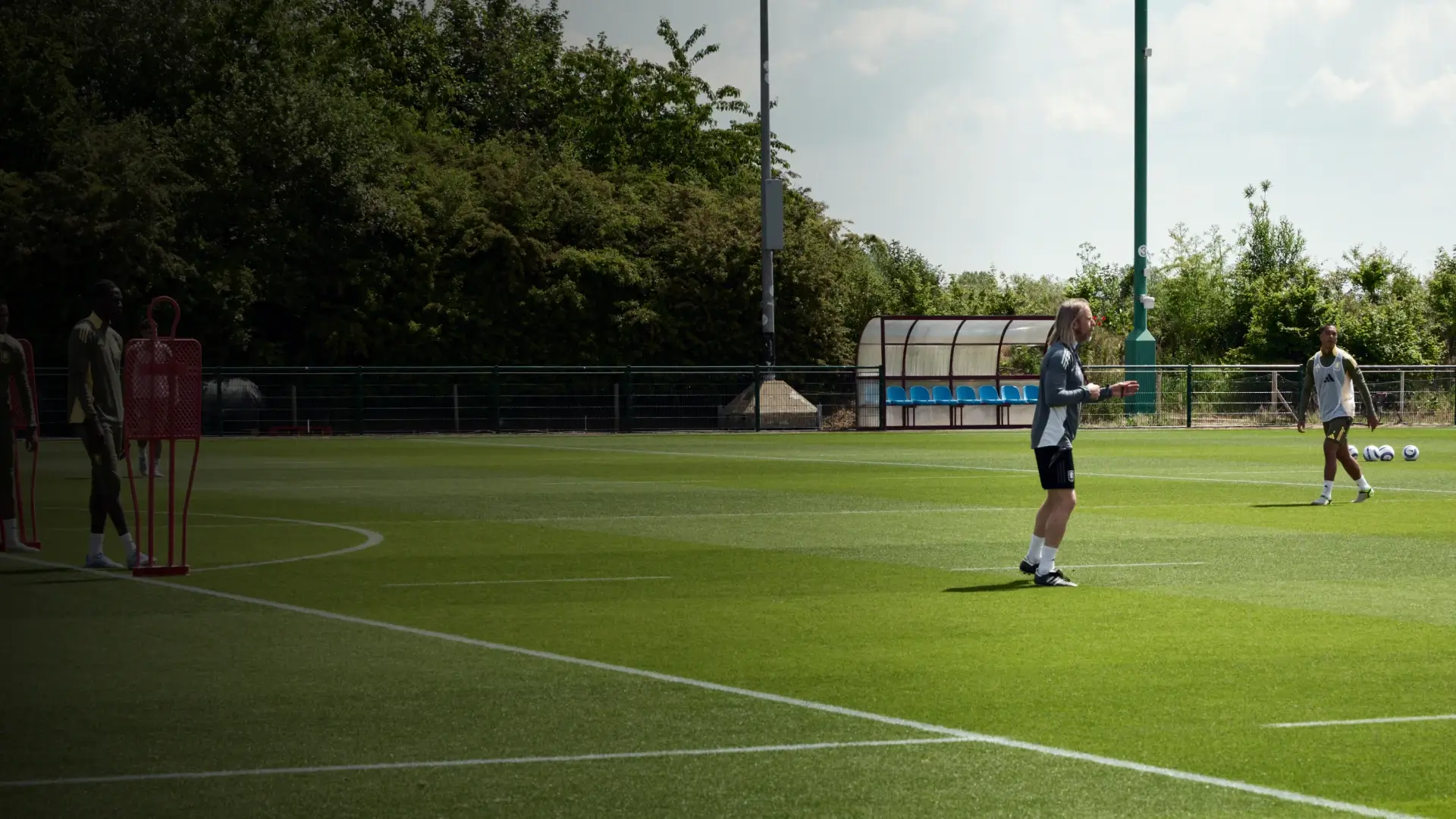 A soccer player trains alone on a lush green field, with a bench and trees in the background under a partly cloudy sky.