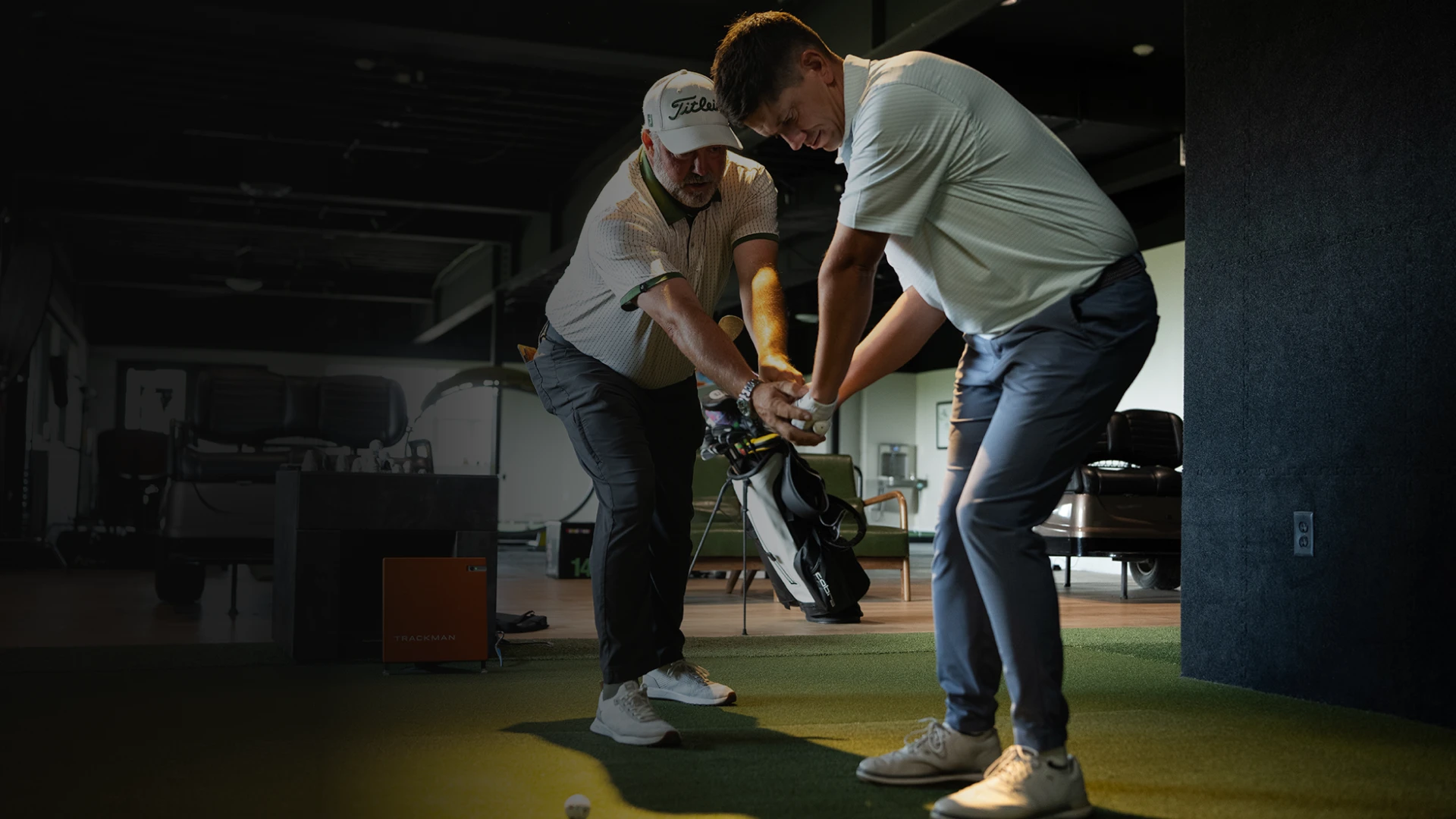 Golf instructor assists a student with swing technique indoors, adjusting grip and posture on a green mat.