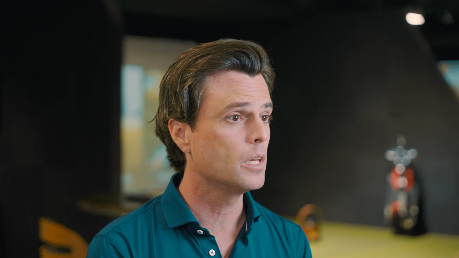 Man in a teal collared shirt speaks inside a dimly lit room with blurred background.