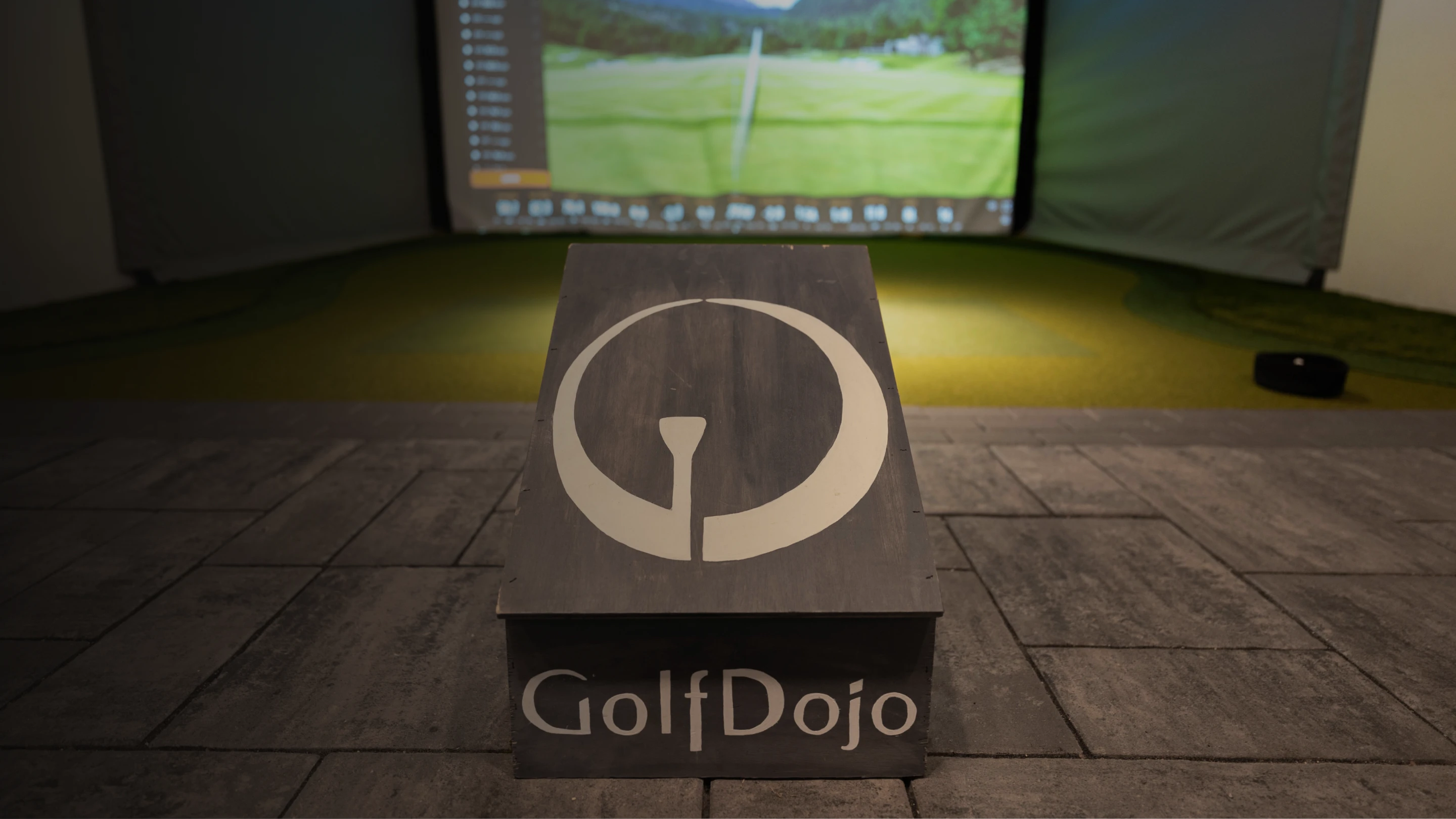 A wooden podium with the logo and text "GolfDojo" stands in front of a golf simulator screen displaying a grassy course.