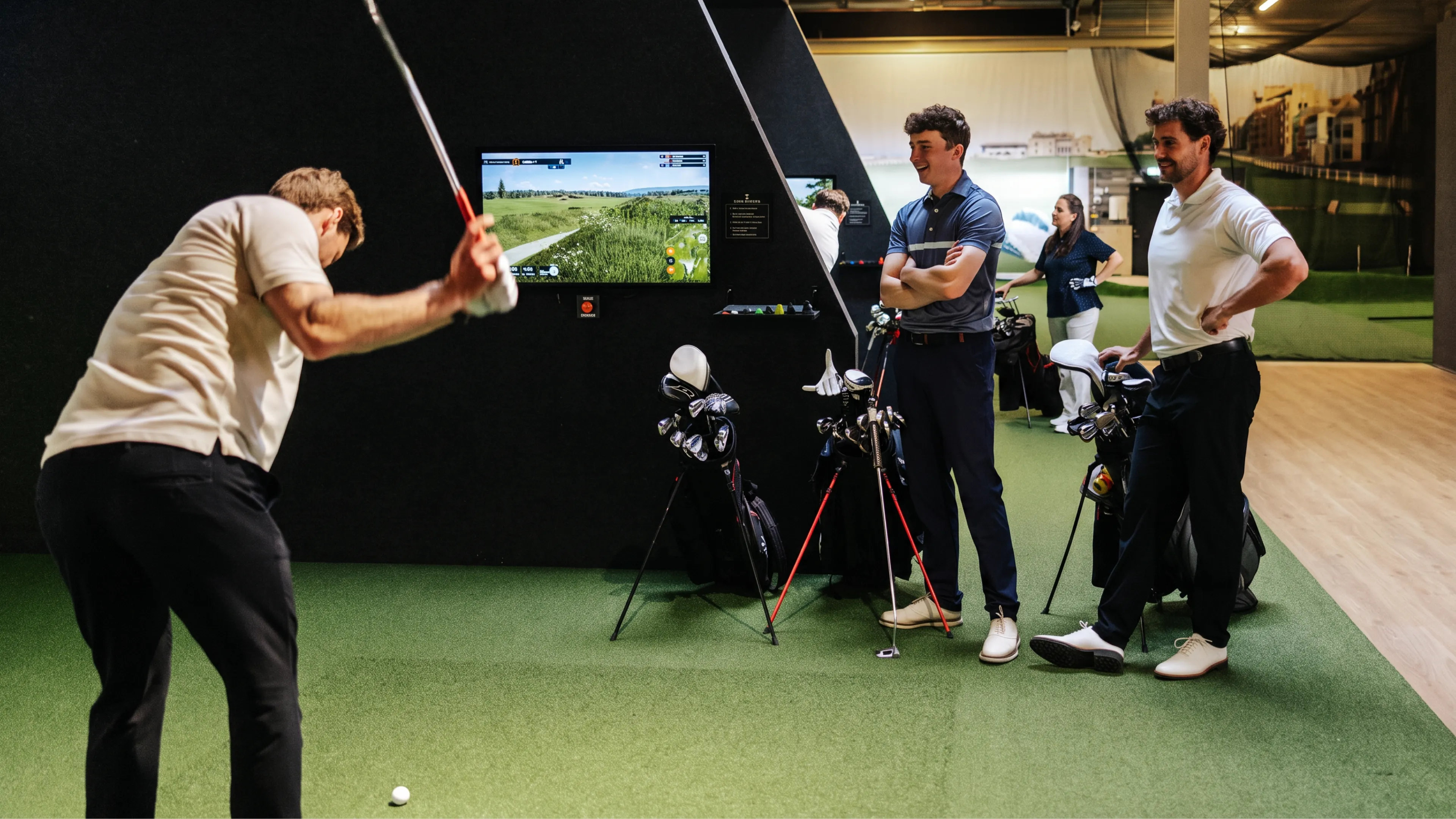 A man swings a golf club indoors while two others watch near a screen showing a virtual golf course; golf bags are visible in the foreground.