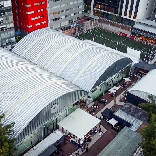 Aerial view of a complex with arched metallic roofs, an adjacent fenced sports field, and surrounding urban buildings.