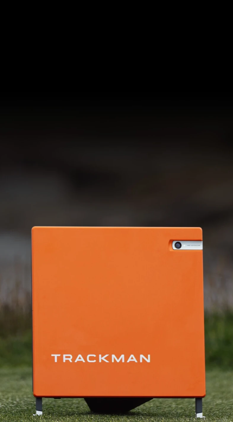 Orange TrackMan device with a small camera on a stand, set against a blurred dark background.