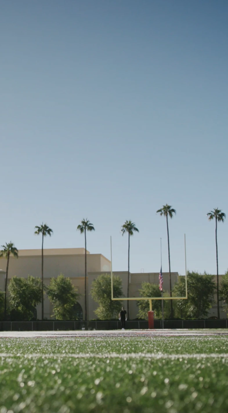 Low-angle view of a football field turf with a yellow goalpost and American flag, a lone person near the post, palm trees and a building.