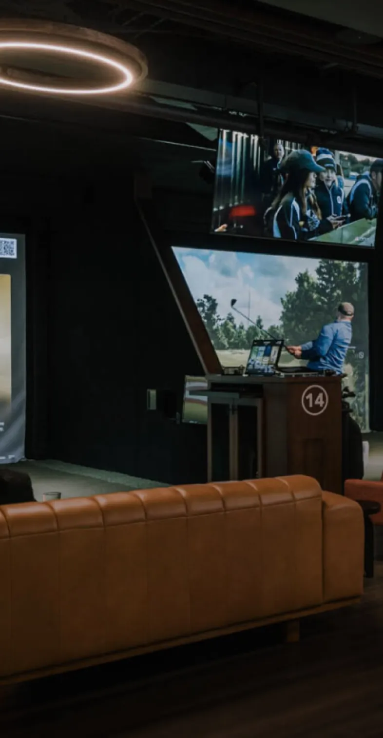 A man plays a golf simulator in a dimly lit room with a leather couch and multiple screens displaying sports.