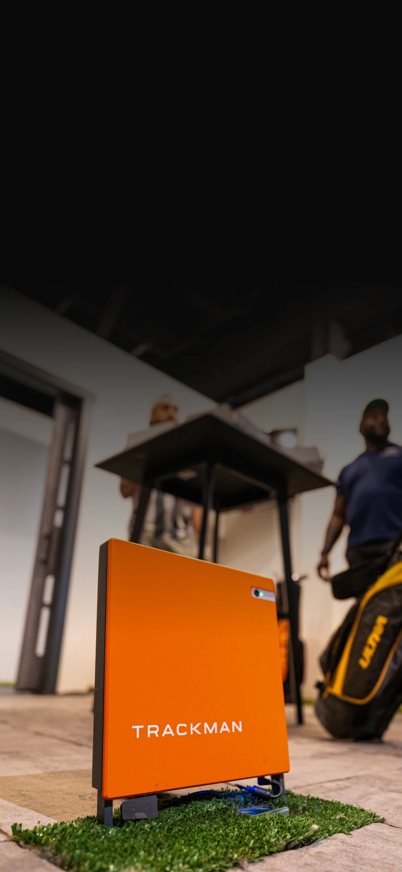 Orange TrackMan device on artificial grass indoors, with two people standing in the background next to a table.