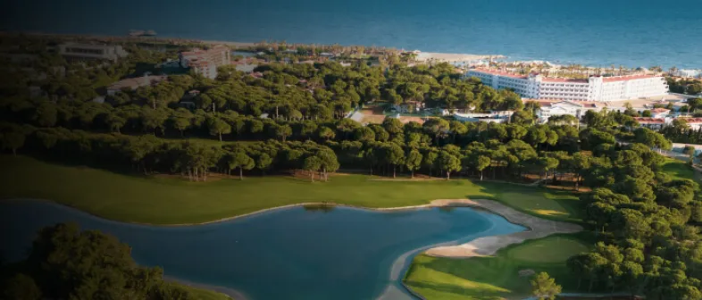 Aerial view of a coastal resort with lush greenery, a golf course, and a lake, adjacent to a sandy beach and blue ocean.
