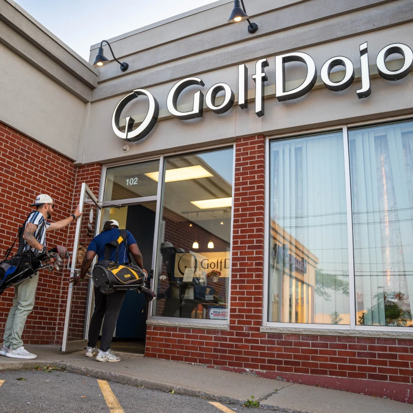 Two people with golf bags enter a building with a sign reading "Golf Dojo" on a brick facade.